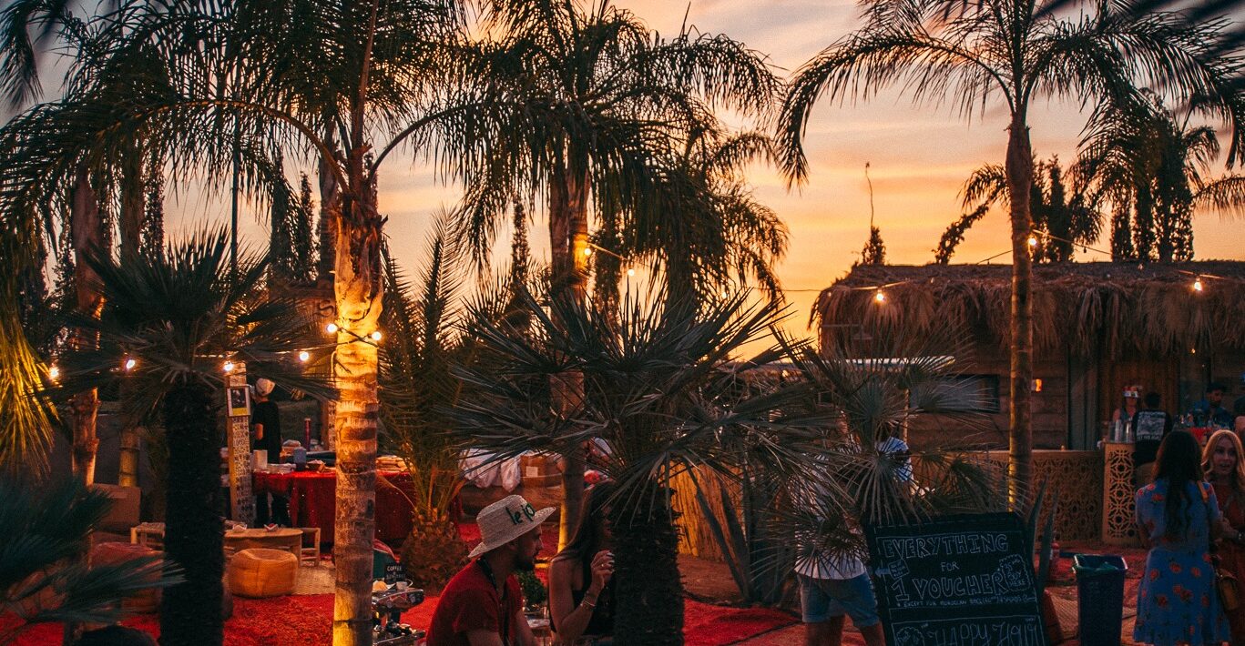 A sunset falls over the Fellah Hotel. The silouhettes of palm trees are illuminated by strings of fairy lights between the trunks.